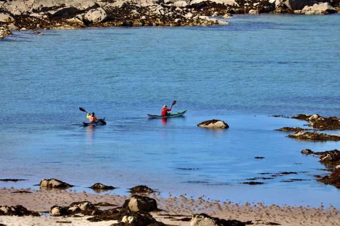 Summer Isles Two kayakers paddling in calm blue water near rocky shoreline.